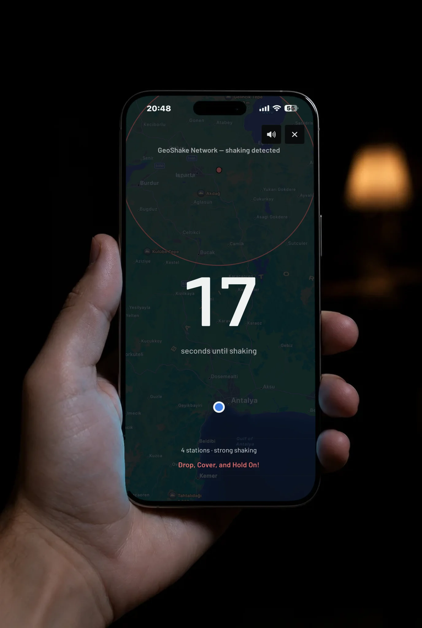 A hand holding a phone at night. On the screen the GeoShake app shows an earthquake alert over a map of Antalya: 17 seconds until shaking, four stations reporting strong shaking, and the instruction to drop, cover and hold on.